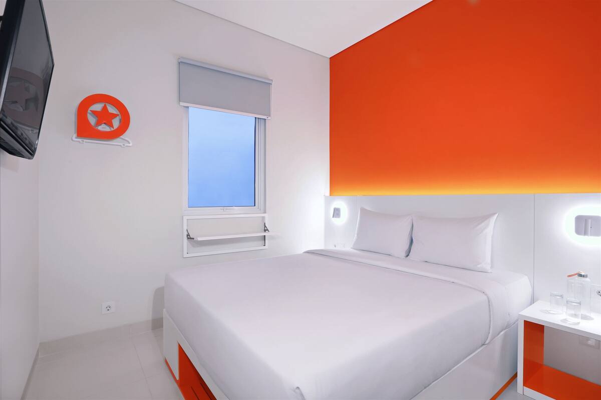 Une chambre d’hôtel moderne avec un grand lit, une fenêtre offrant une belle vue, un téléviseur fixé au mur et un mur d’accent de couleur orange vif.