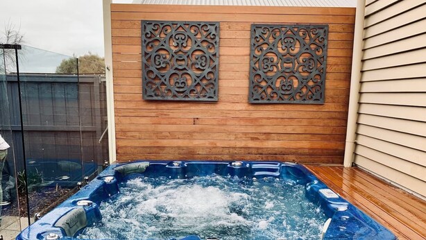 Un jacuzzi en una terraza de madera, con un muro de madera y paneles metálicos decorativos al fondo.