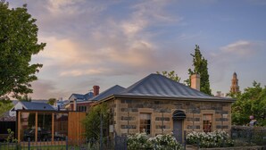 A stone building with a pitched roof, a wooden porch, and a small garden with white flowers. - Fusilier Cottage Battery Point (Battery Point)