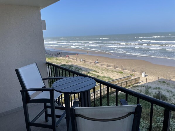 A balcony with a table and chairs overlooking a beach with a wooden fence and a boardwalk.