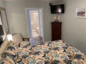 A bedroom with a bed, a chair, a TV, and a door leading to a bathroom.