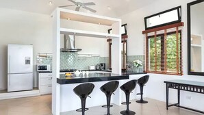 A modern kitchen with a central island, three bar stools, a refrigerator, and a window with blinds. - Luxury Beachfront Tai Pan Villa (Wichit)
