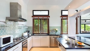 A modern kitchen with a microwave, stove, sink, and dishwasher. There are two windows with blinds, a large window with a view of greenery, and a countertop with a fruit bowl and teapot. - Luxury Beachfront Tai Pan Villa (Wichit)