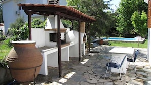 Un patio al aire libre con suelo de piedra, una maceta terracota grande, una parrilla y una piscina al fondo.