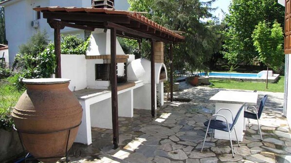 An outdoor patio with a stone floor, a large terracotta pot, a grill, and a pool in the background.