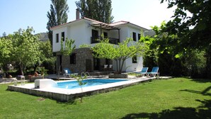 Una casa de dos pisos con piscina, fachada de piedra y jardín con árboles y sillones.