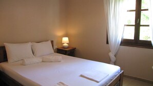 Una cama bien hecha con sábanas y almohadas blancas, una lámpara de noche y una ventana con cortinas.