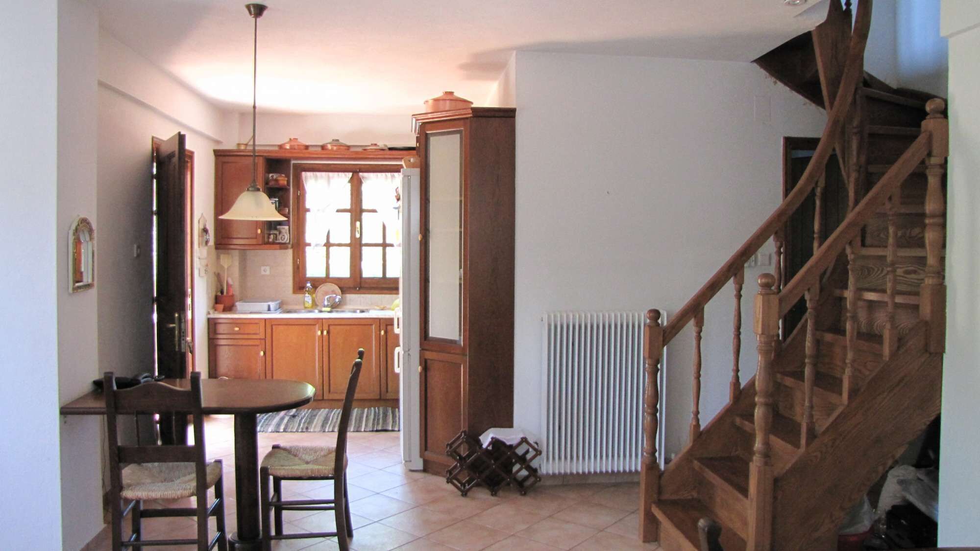 A kitchen with wooden cabinets, a dining table with chairs, a hanging light fixture, and a staircase.