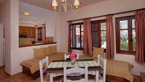 Ein gemütliches Zimmer mit Esstisch, Stühlen, einer Bank, einer Chandelüster, einer Küchenzeile und großen, mit Vorhängen versehenen Fenstern.