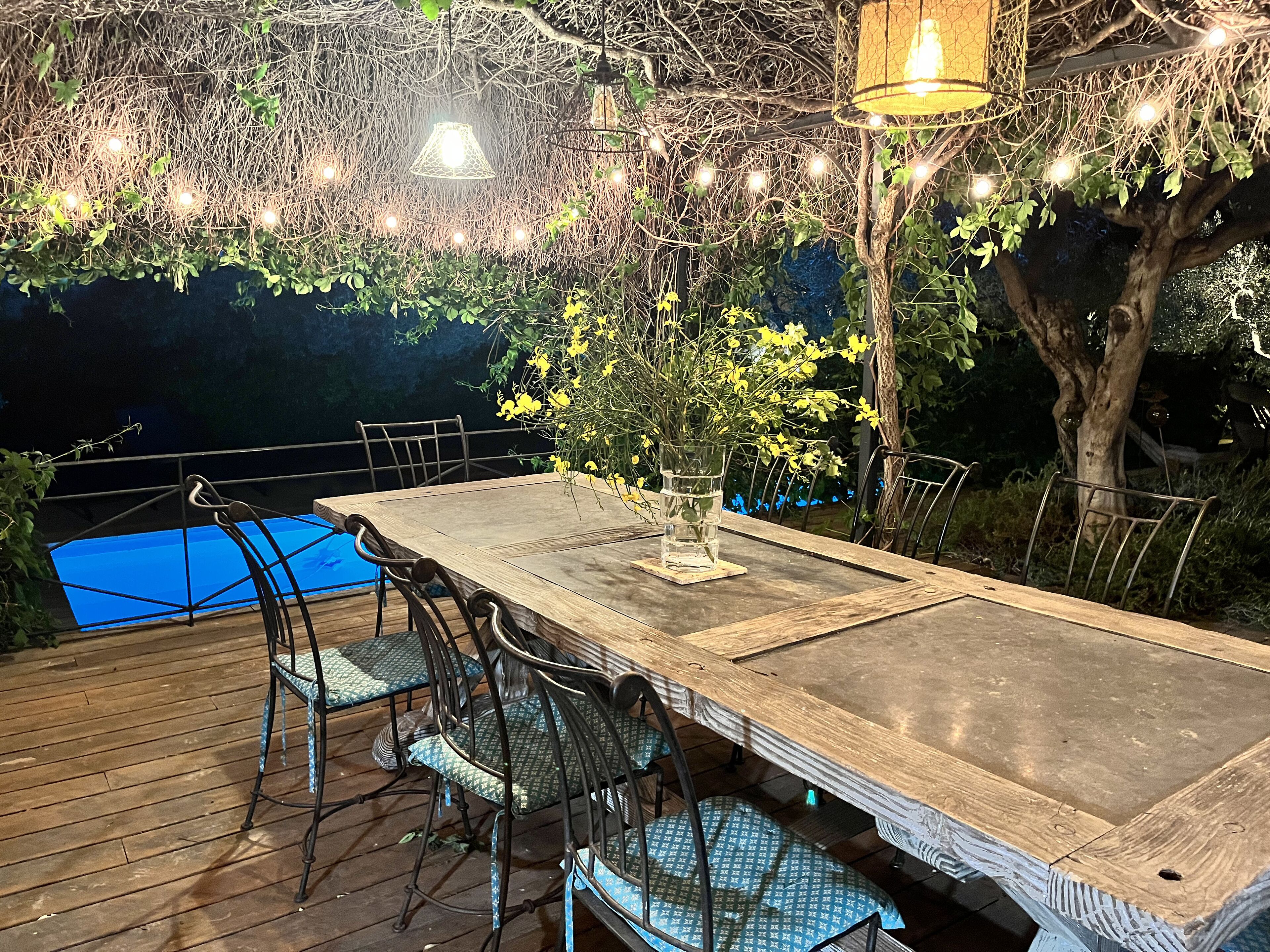 An outdoor dining area with a wooden table and metal chairs, surrounded by greenery and string lights.