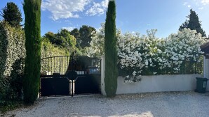 A gravel driveway leading to a gated entrance with a white wall and flowering plants. - Belle Villa au Coeur des Alpilles (Maussane-les-Alpilles)