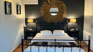 A modern bedroom with a large bed, two bedside tables with lamps, a wall-mounted air conditioner, and framed pictures on the wall. - Belle Villa au Coeur des Alpilles (Maussane-les-Alpilles)