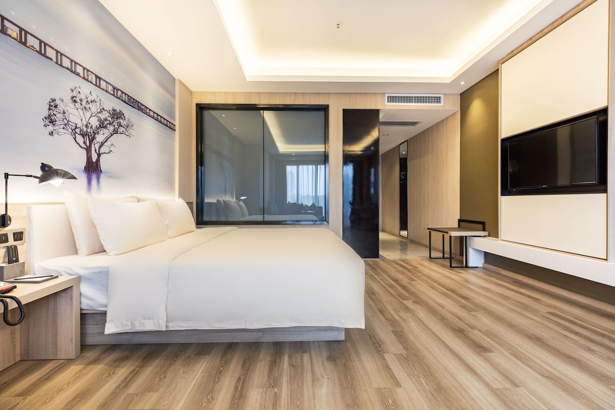 Ein modernes Hotelzimmer mit einem großen Bett, einem Fernseher, einem Schreibtisch mit Lampe und einem großen Fenster.