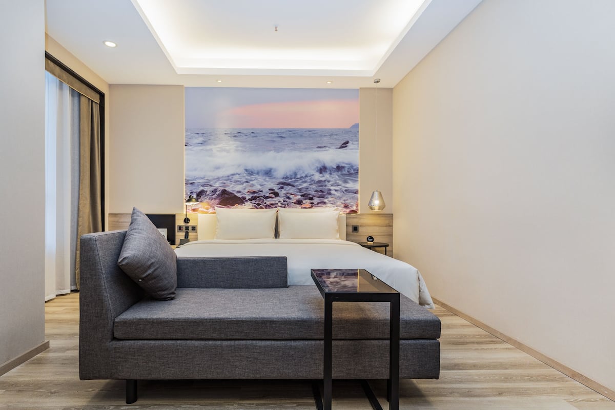 Ein modernes Schlafzimmer mit einem großen Bett, einem grauen Sofa, einem kleinen Tisch und einer Wandmalerei mit einer Strandansicht.