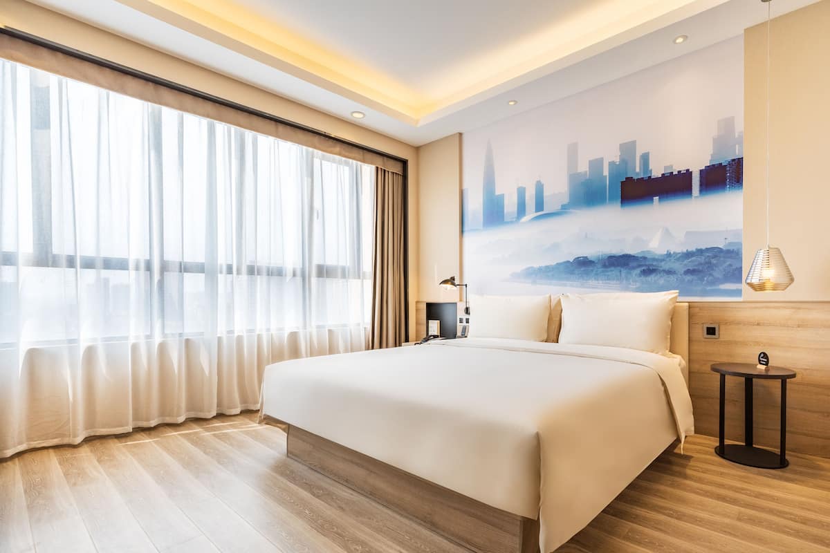 Ein Hotelzimmer mit einem großen Bett, einem Stadtbild-Wandgemälde, einem Fenster mit transparenten Vorhängen und einem Holzboden.