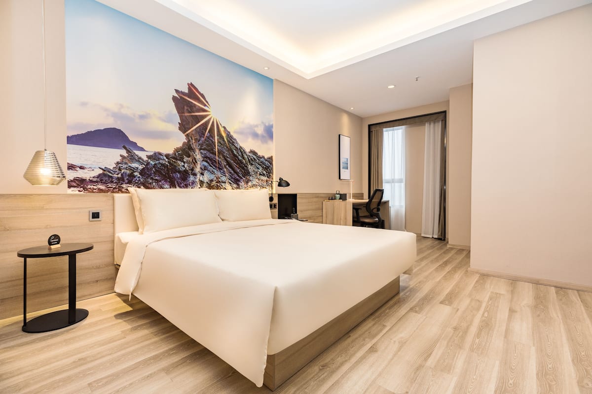 Ein modernes Hotelzimmer mit einem großen Bett, einem Schreibtisch, einem Sessel, einem kleinen Tisch und einem Gemälde einer Küstenlandschaft.