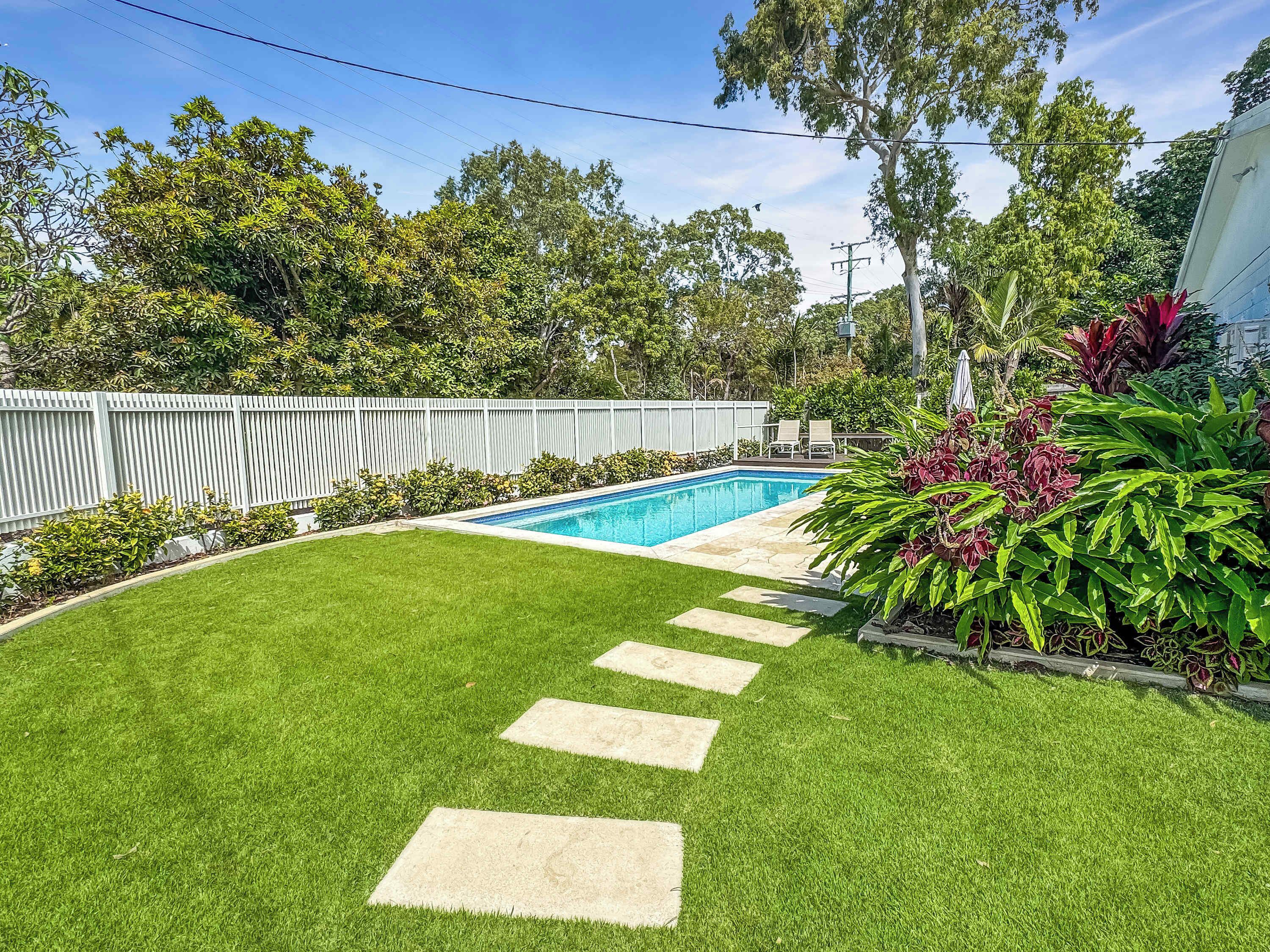 A backyard with a swimming pool, a white fence, and a well-maintained lawn.