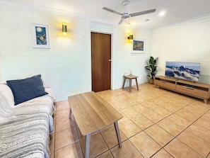 A living room with a sofa, a wooden table, a TV on a wooden stand, a ceiling fan, and a door.