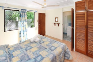 Une chambre à coucher avec un lit, un ventilateur de plafond, une fenêtre avec des rideaux, une porte en bois et une salle de bain visible à travers une porte ouverte.
