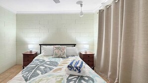 Un dormitorio con cama, dos mesitas de noche, ventilador de techo y ventana con cortinas.