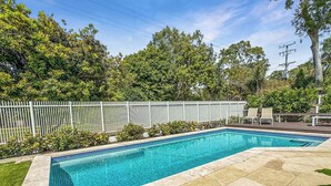 Una piscina rectangular con borde de azulejos, rodeada por una cerca blanca y abundante vegetación.
