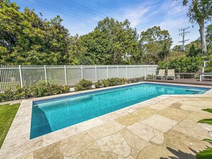 A rectangular swimming pool with a tiled edge, surrounded by a white fence and lush greenery.