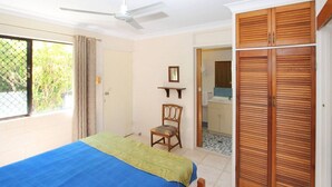 Une chambre à coucher comprenant un lit, une chaise, une armoire, un ventilateur de plafond, et une salle de bain visible à travers une porte ouverte.