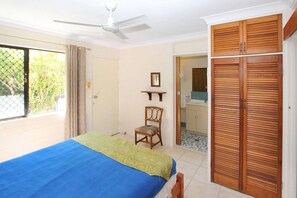 Un dormitorio con cama, una silla, un armario, un ventilador de techo y un baño visible a través de una puerta abierta.