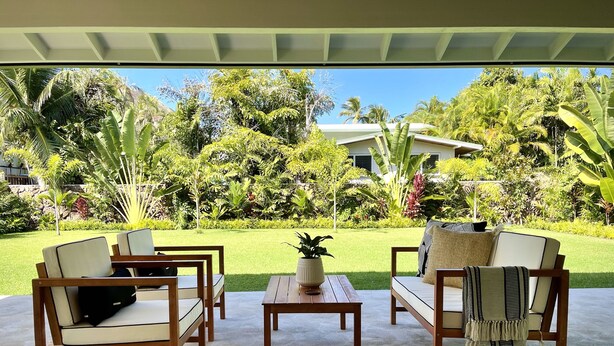 Un patio con mobiliario de madera, una mesa central con planta en maceta, y vistas a un jardín exuberante con plantas tropicales variadas y un edificio al fondo.