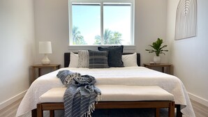 Un dormitorio ordenado con una cama grande, una mesita de noche con lámpara, una planta en maceta y vistas a palmeras a través de la ventana.
