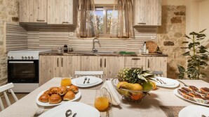 A kitchen with wooden cabinets, a window with curtains, a sink, and a dining table set with food and drinks. - Poseidon Apartments (Platanias)