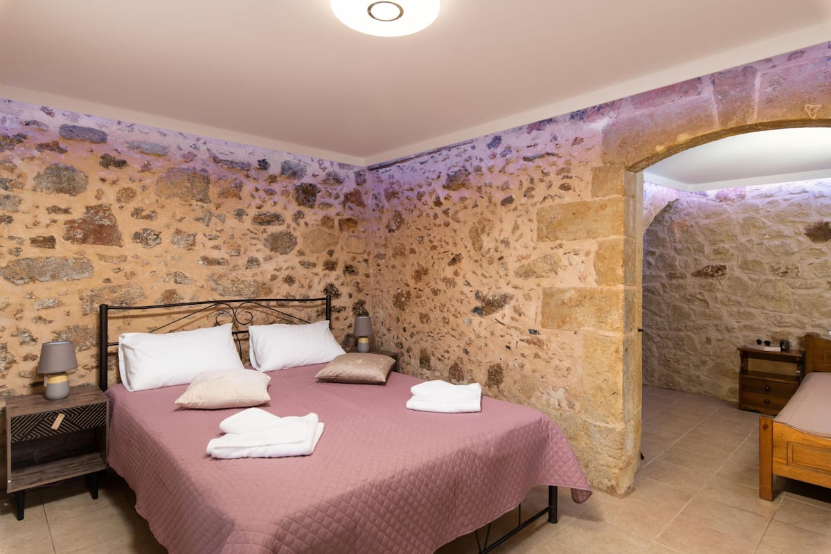 A bedroom with a stone wall, a bed with a pink quilt, and a wooden nightstand with a lamp.