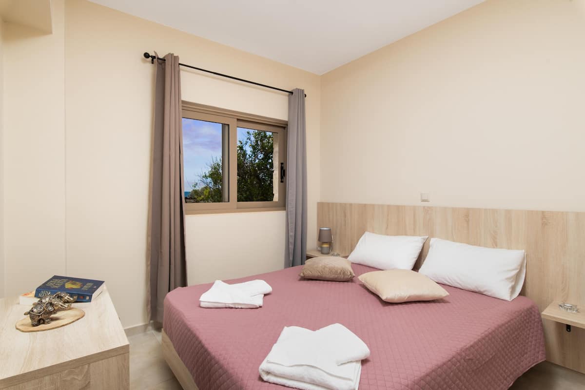 A neatly made bed with a pink bedspread, white pillows, and a window with curtains. There is a bedside table with a lamp, a book, and a decorative item.