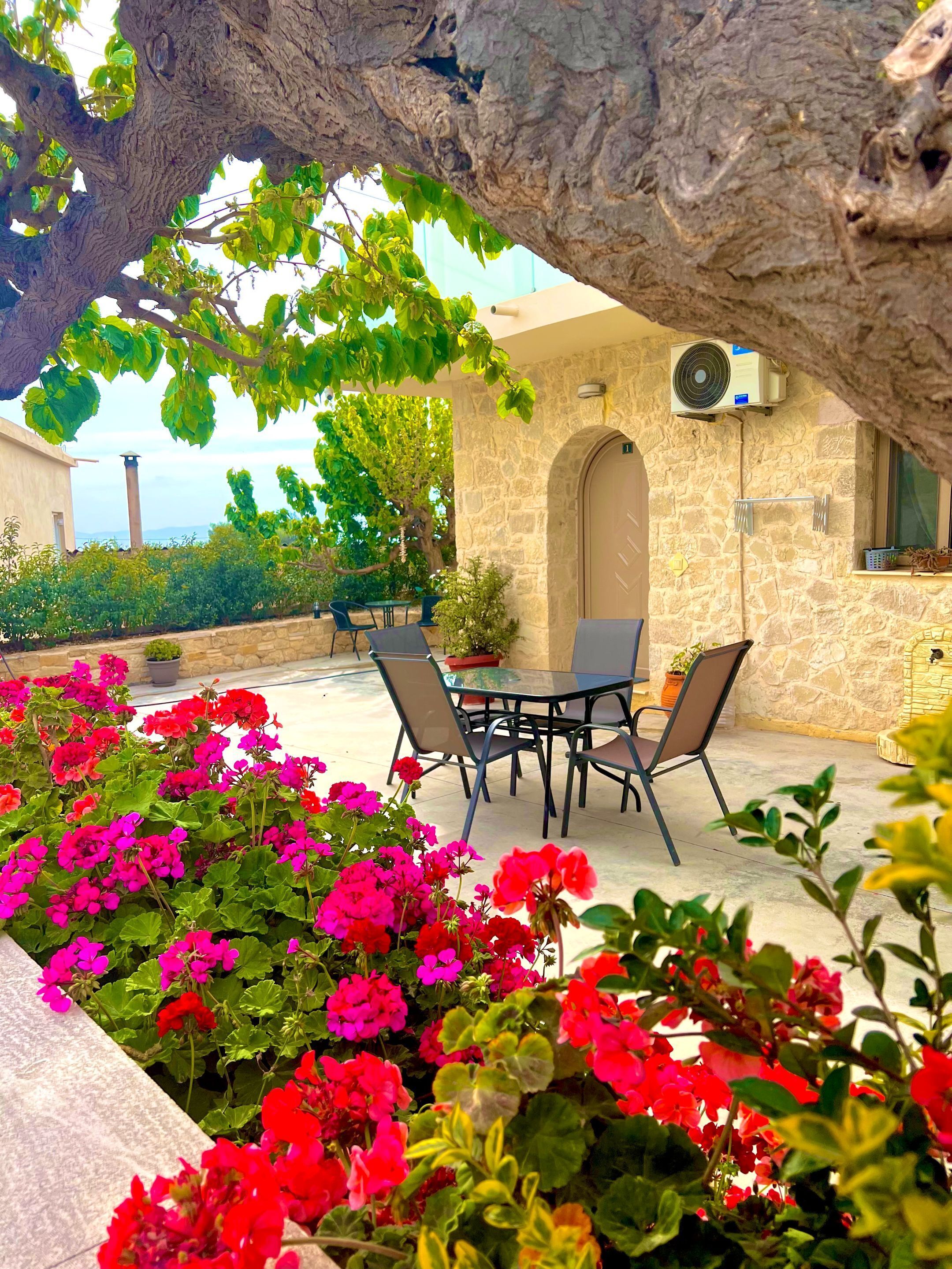 Ein Terrassenbereich im Freien mit einer Steinsäule, einem Tisch mit Stühlen und leuchtenden Blumen.