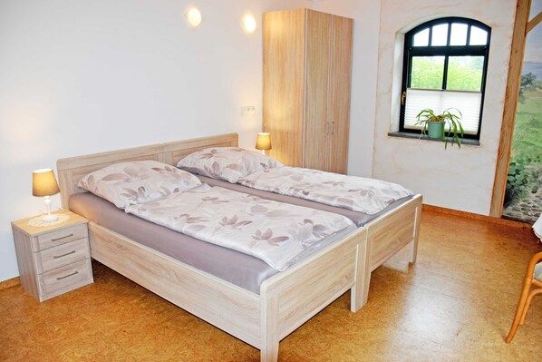 A bedroom with a bed, bedside tables, a wardrobe, a window with a plant, and a view of greenery.
