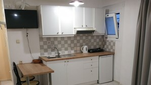 A compact hotel room with a bed, a small dining table with chairs, a kitchenette, and a TV mounted on the wall.