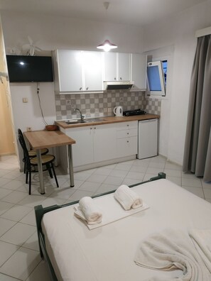 A compact hotel room with a bed, a small dining table with chairs, a kitchenette, and a TV mounted on the wall. - Renia Studios Economy Studio Ground Floor (Malevizi)