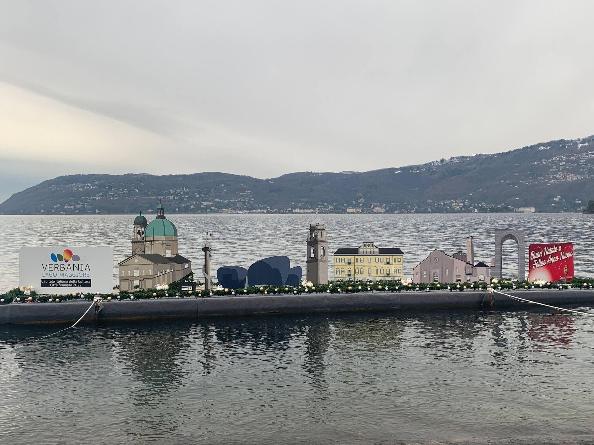 Vista al frente del lago con un cartel de Verbania Lago Maggiore, un edificio con cúpula verde y un cartel rojo con texto en italiano.