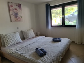 Ein Schlafzimmer mit Bett, Kissen, Decke und einem Fenster mit Blick auf ein Auto und Grünfläche draußen.