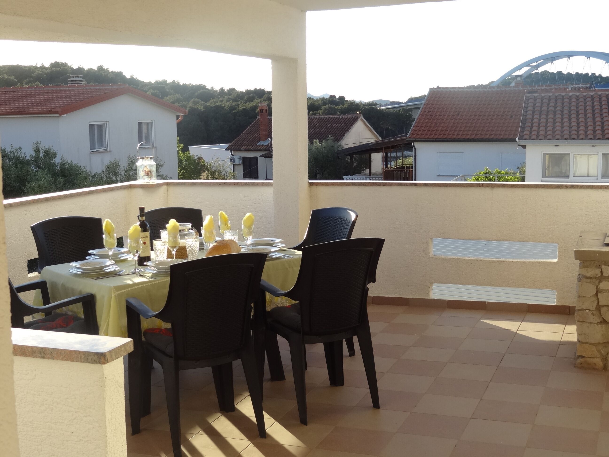 A terrace with a table set for dining, surrounded by chairs, with a view of houses and a hill in the background.