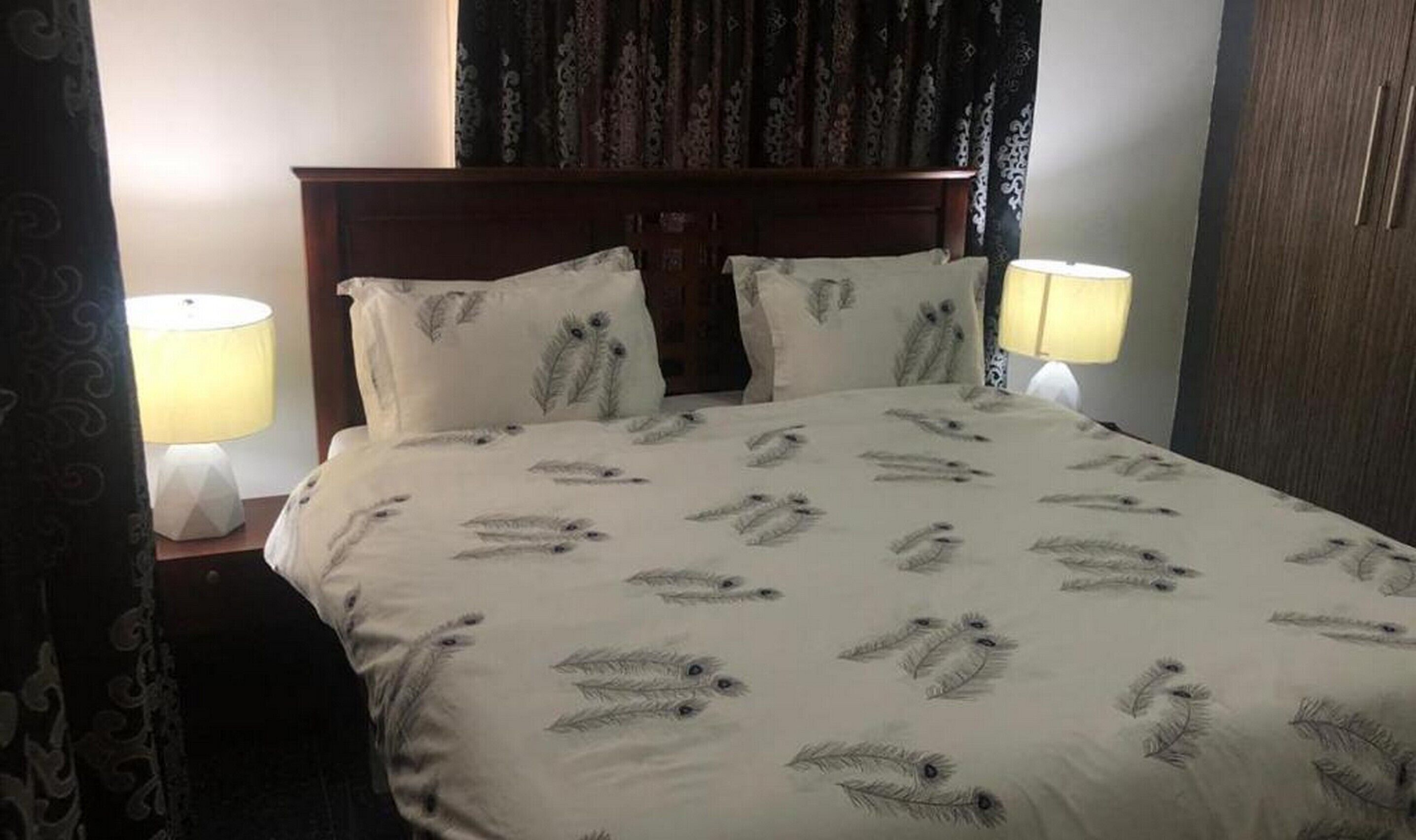 A hotel room with a bed featuring a feather pattern, two bedside lamps, and a dark curtain.