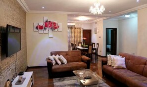 A living room with two brown leather sofas, a coffee table, a TV, and a chandelier. - St Georgess Height-room With Balcony (Nsawam)