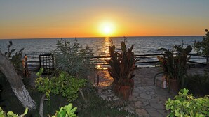 Une vue sur le coucher de soleil au-dessus de l’océan, avec un sentier de pierres menant à une porte, le tout entouré de plantes en pot.