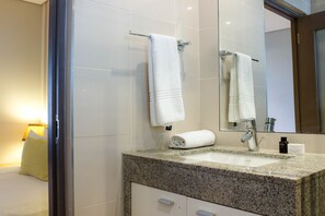 Ein Badezimmer mit einer Granitplatte, einem weißen Waschbecken und einem Spiegel. Zwei Handtücher hängen an der Wand und auf der Platte steht eine kleine Flasche.