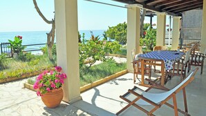 A covered outdoor seating area with a table and chairs, overlooking a garden and the sea.