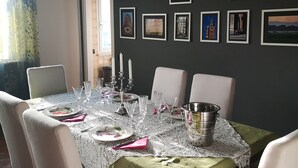 A dining area with a table set for four, white chairs, a lace tablecloth, and a green runner. The wall features framed pictures and a sign reading "VILLA PIERA 1946". - Room in Apartment - Villa Piera Holiday Home in Cremona Apartment With Independent Entrance (Cremona)