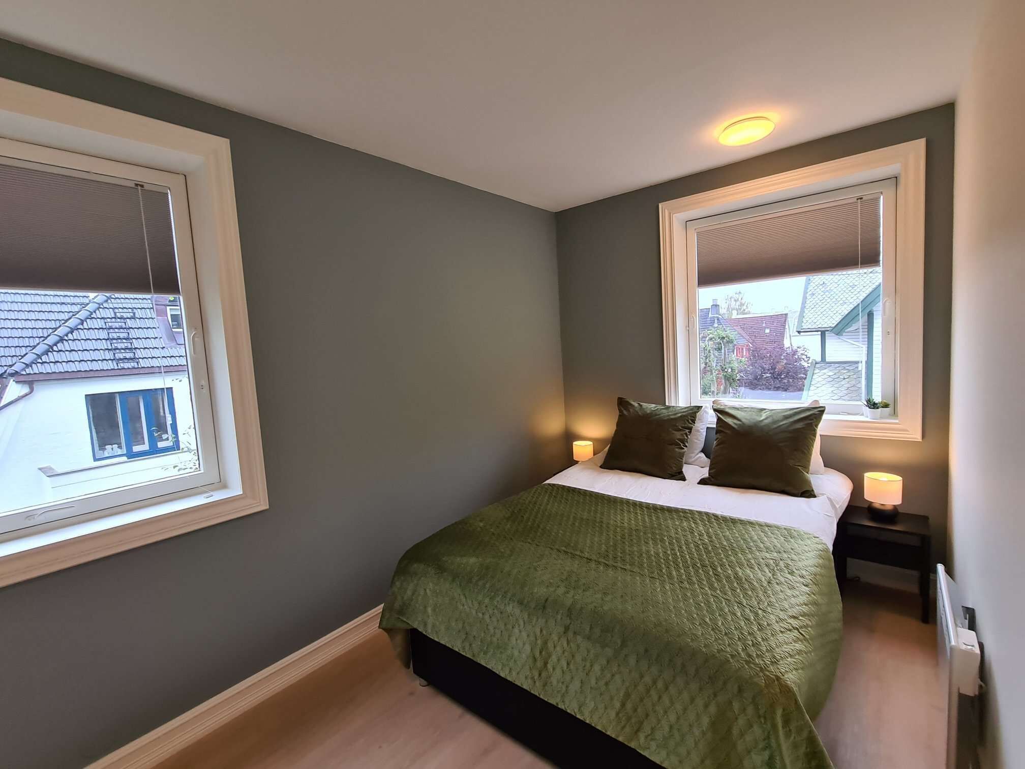 A bedroom with a bed, two bedside tables, two lamps, and two windows.