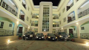Een meerlaags hotel met een centraal atrium, meerdere balkons en een parkeerplaats met verschillende auto's.