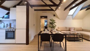 Un appartement moderne de style « loft », avec des poutres apparentes, une cuisine dotée d’un four encastré, un coin repas avec une table dressée pour quatre personnes et un salon avec un canapé et une table basse.