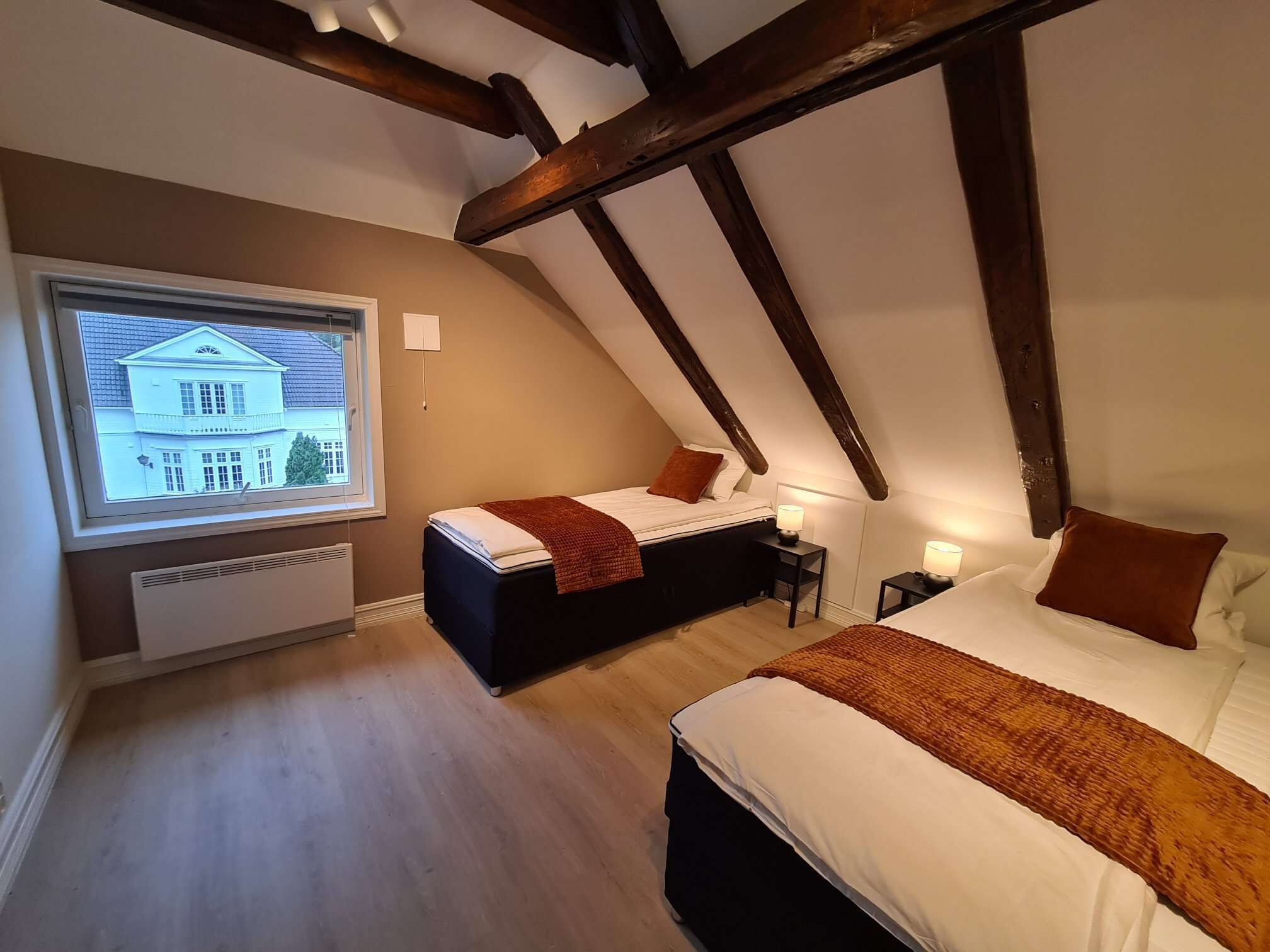 A bedroom with a sloped ceiling, a large window with a view of a house, two beds with brown bedding, and a heater under the window.
